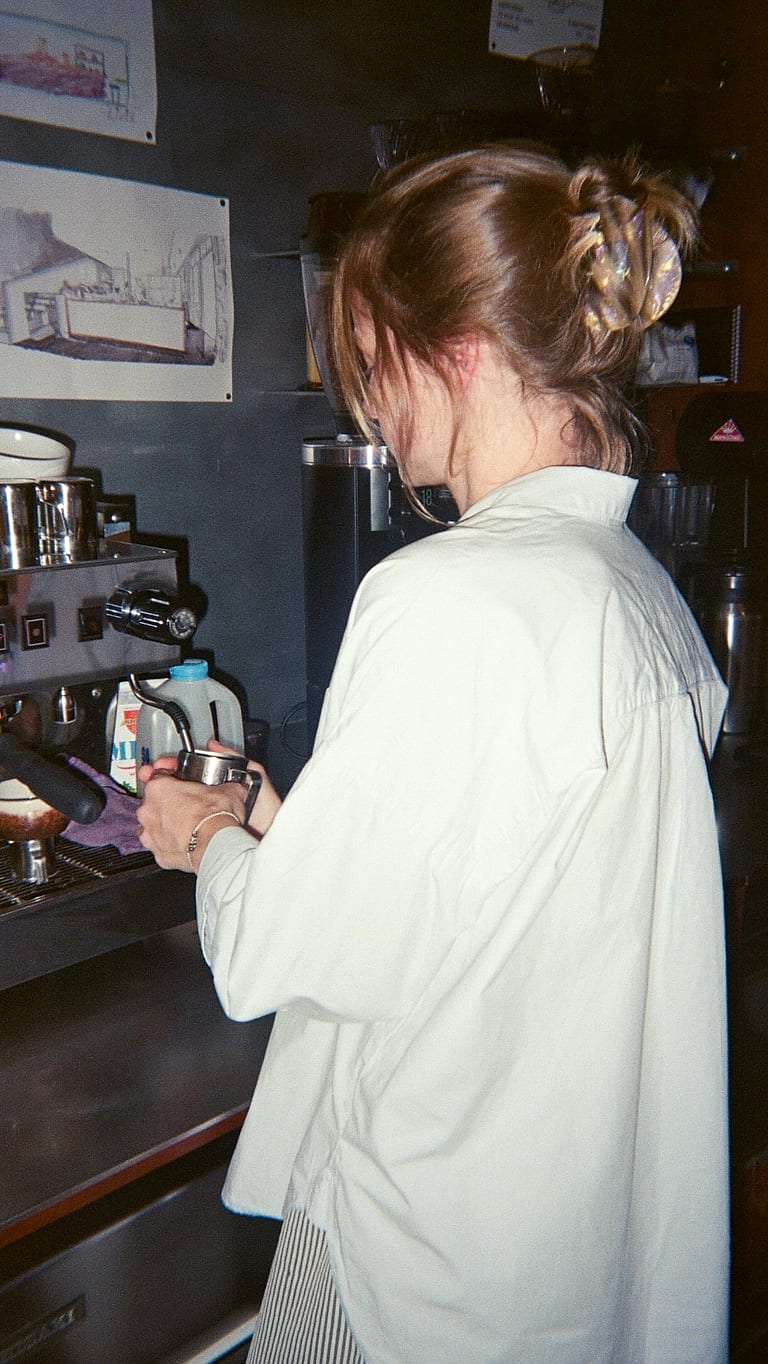 Au comptoir, de dos, en chemise blanche, devant la machine à espresso.