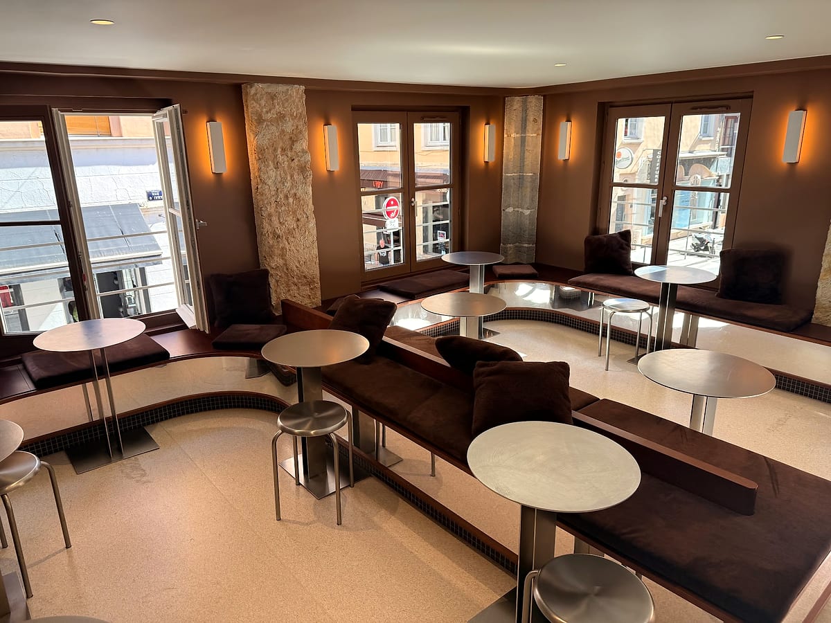 La banquette de velours brun et les tables rondes de l'étage, dans la lumière des fenêtres d'angle.