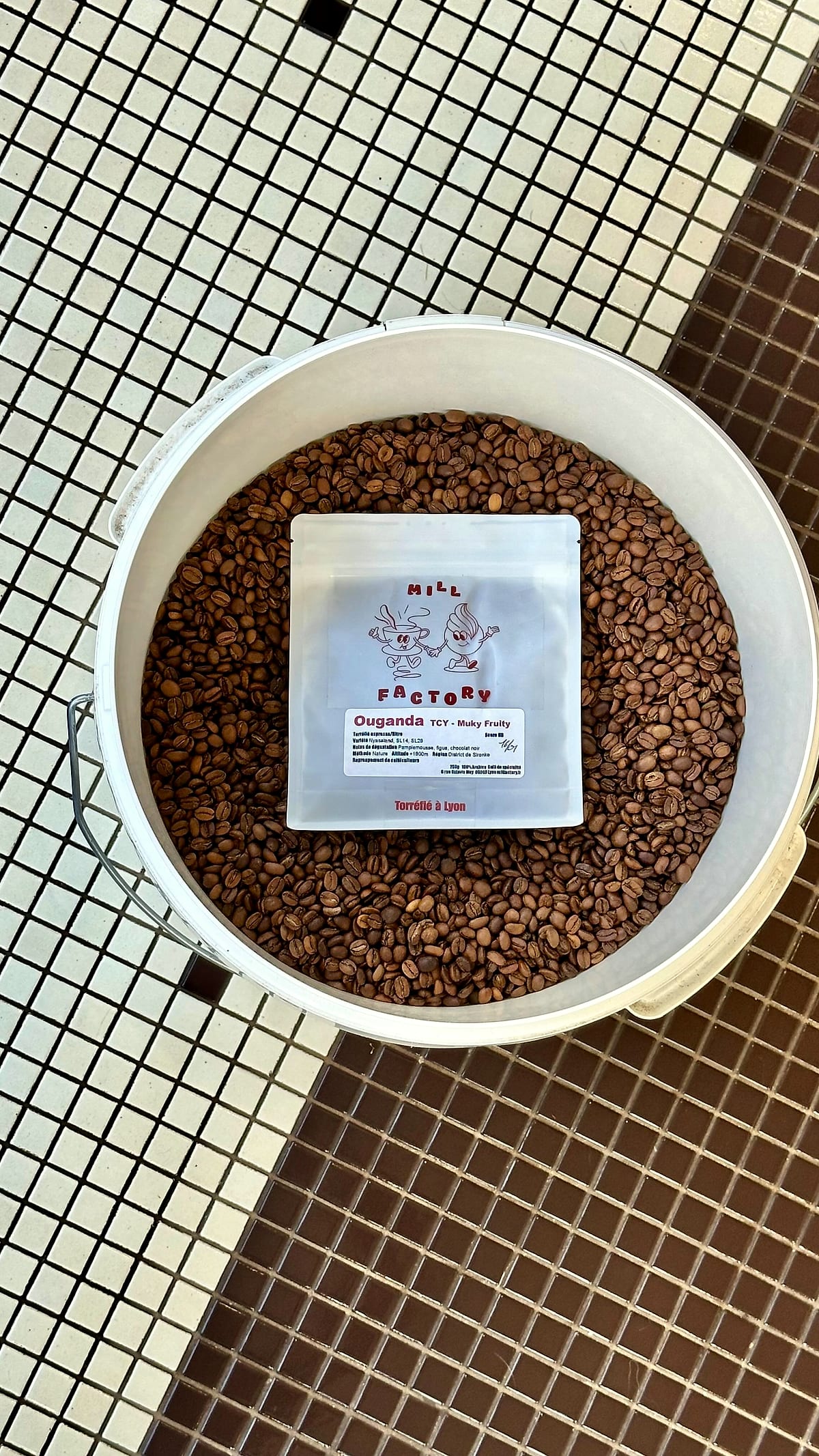 Un seau de grains et un sachet Mill Factory venu d'Ouganda, posés sur le carrelage mosaïque.
