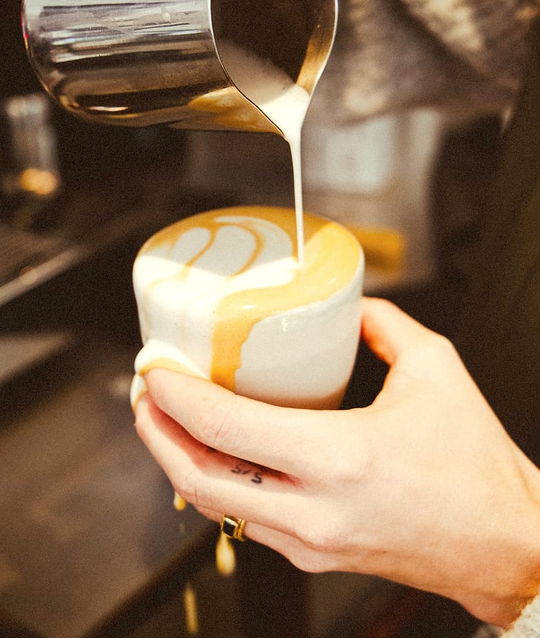 Le lait versé en filet dans la tasse, les mains du barista autour.