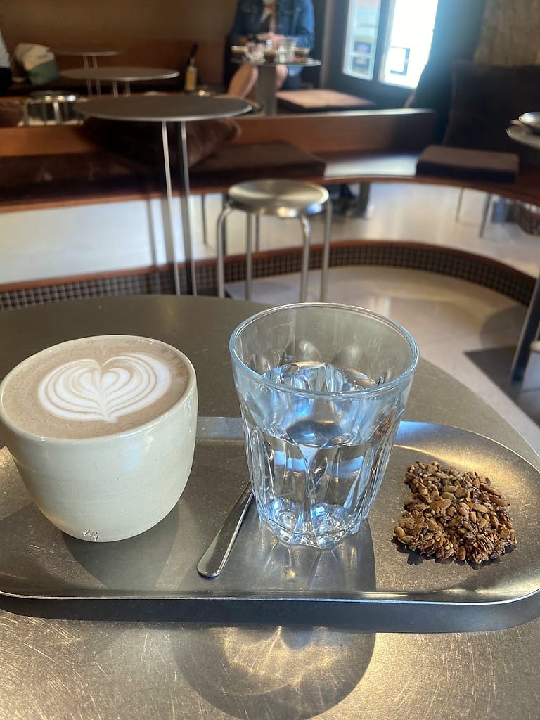 Un cappuccino au motif dessiné, un verre d'eau et un carré de granola, servis sur un plateau en inox.